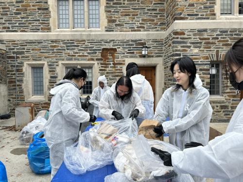 6 students lean over a table and wear protective jumpsuits while sorting trash from recyclables in a waste audit outside a residence hall.