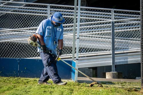 A person wearing a Duke employee uniform and hat edges the lawn with a weed eater landscaping tool. 