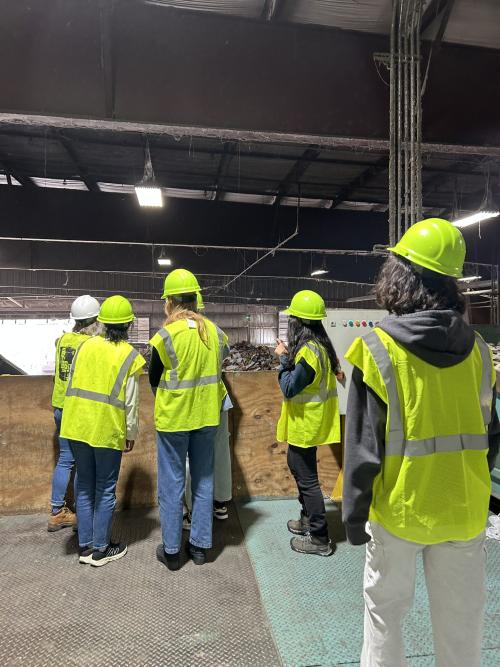 5 people wearing bright yellow personal protective equipment including vests and helmets observe a recycling facility.